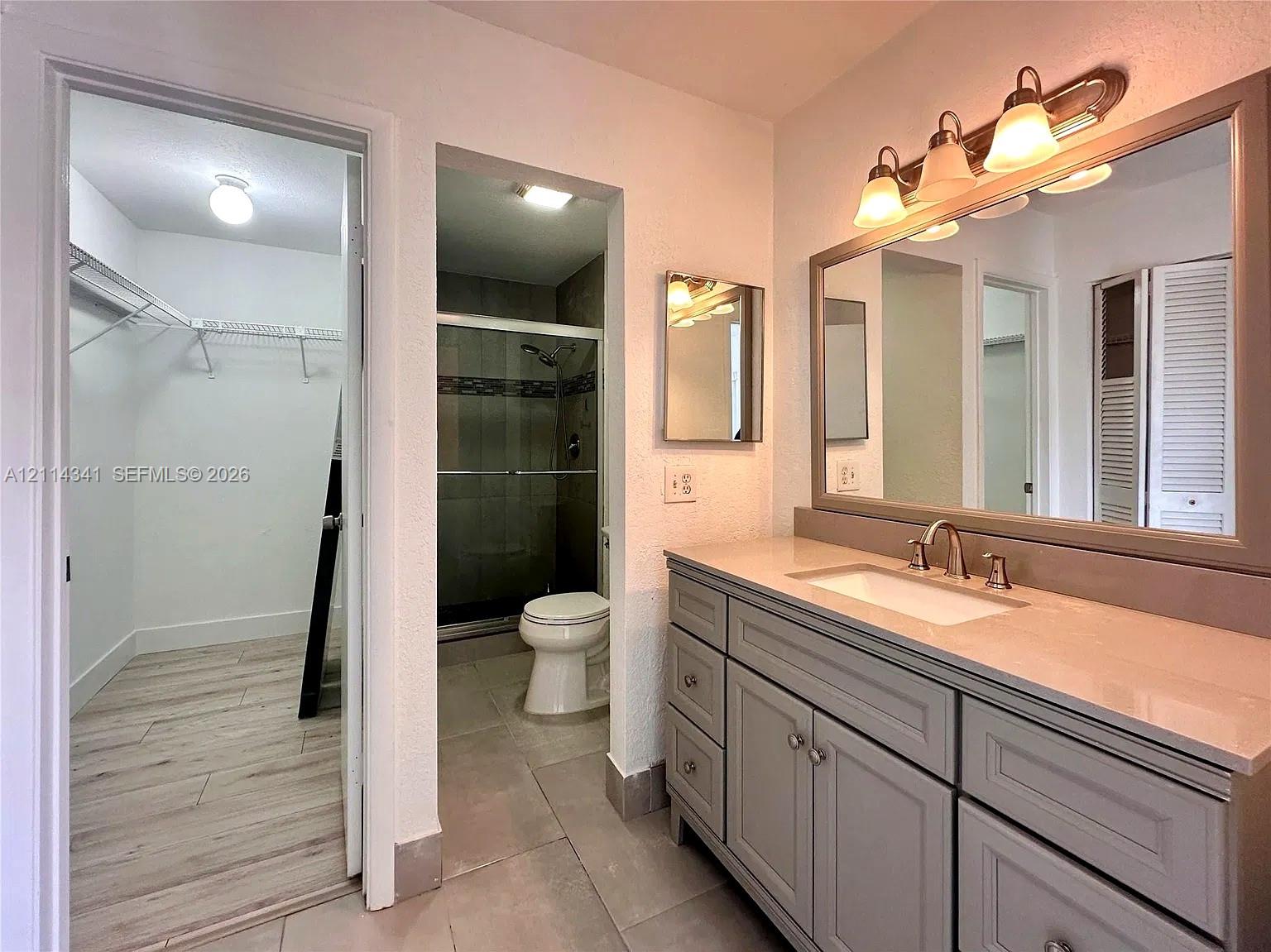 Bathroom featuring a vanity with shaker-style cabinetry, a solid-surface countertop with an integrated sink, a framed mirror, a multi-bulb light fixture, and a walk-in shower with tile surround Bathroom featuring a vanity with shaker-style cabinetry, a solid-surface countertop with an integrated sink, a framed mirror, a multi-bulb light fixture, and a walk-in shower with tile surround