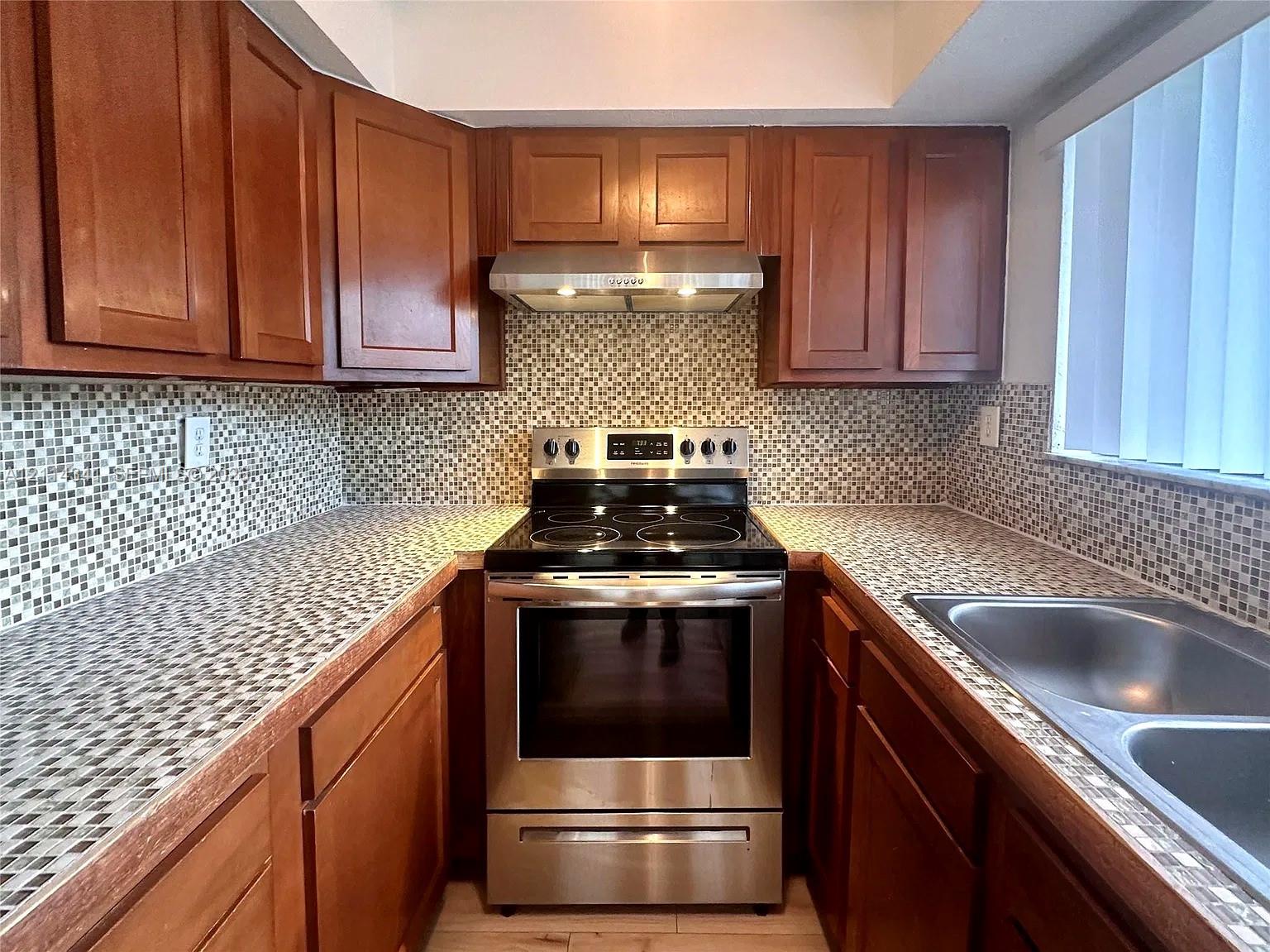 Kitchen featuring wood cabinetry, stainless steel electric range, stainless steel range hood, mosaic tile backsplash, and double-basin stainless steel sink Kitchen featuring wood cabinetry, stainless steel electric range, stainless steel range hood, mosaic tile backsplash, and double-basin stainless steel sink