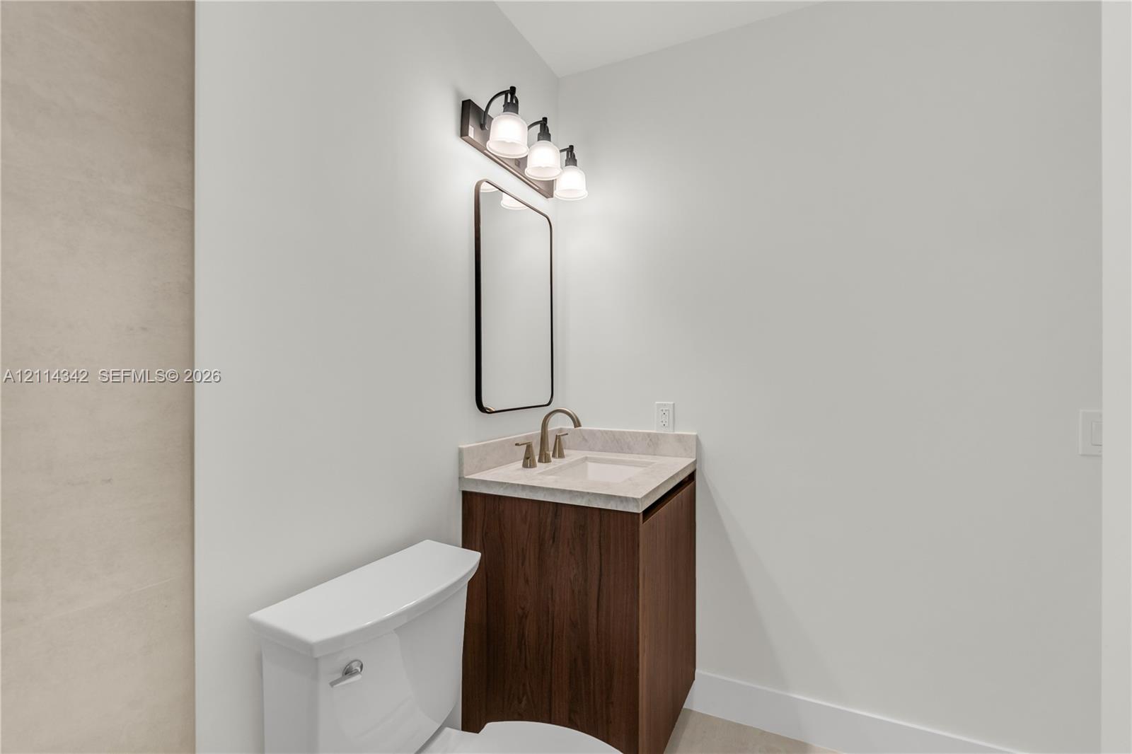 Modern vanity with a neutral stone-top surface and drop-in sink, paired with a dark wood-finish cabinet, rectangular mirror with rounded edges, and a three-bulb light fixture