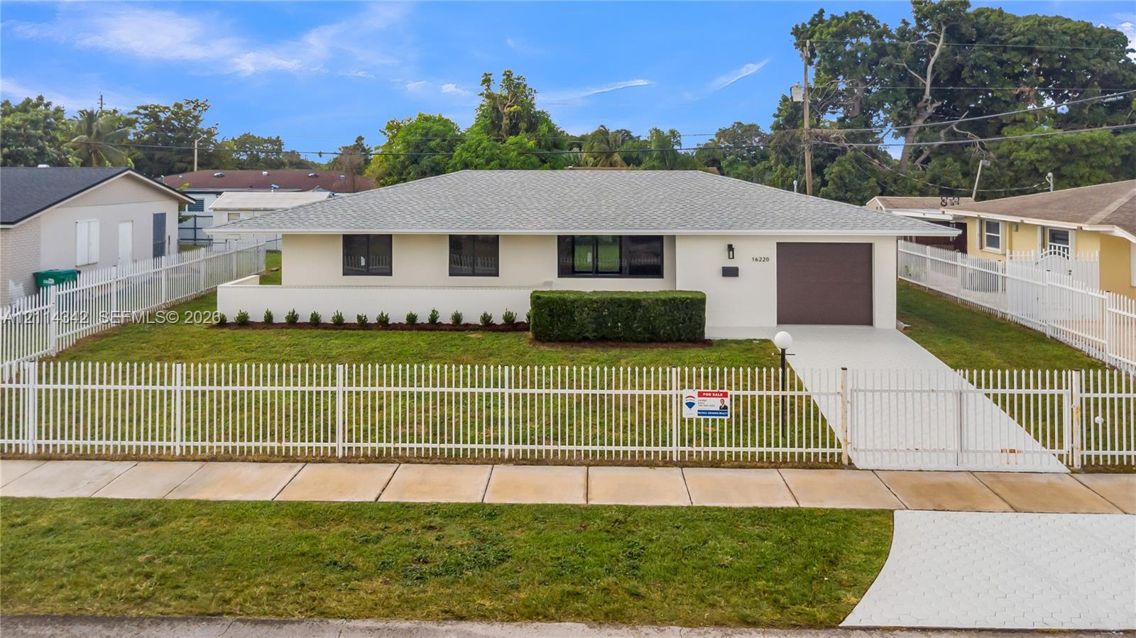 Single-story residence featuring a light-toned exterior, asphalt shingle roof, attached garage, and a full-perimeter white metal fence enclosing the front lawn