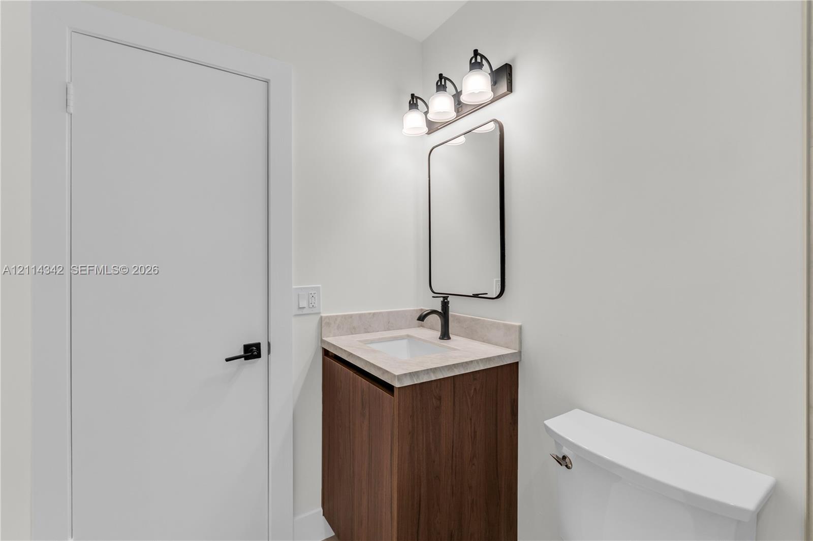 Bathroom featuring a wood-finish vanity with a stone-composite countertop, undermount sink, matte black faucet, and rounded-corner mirror with coordinating three-light fixture
