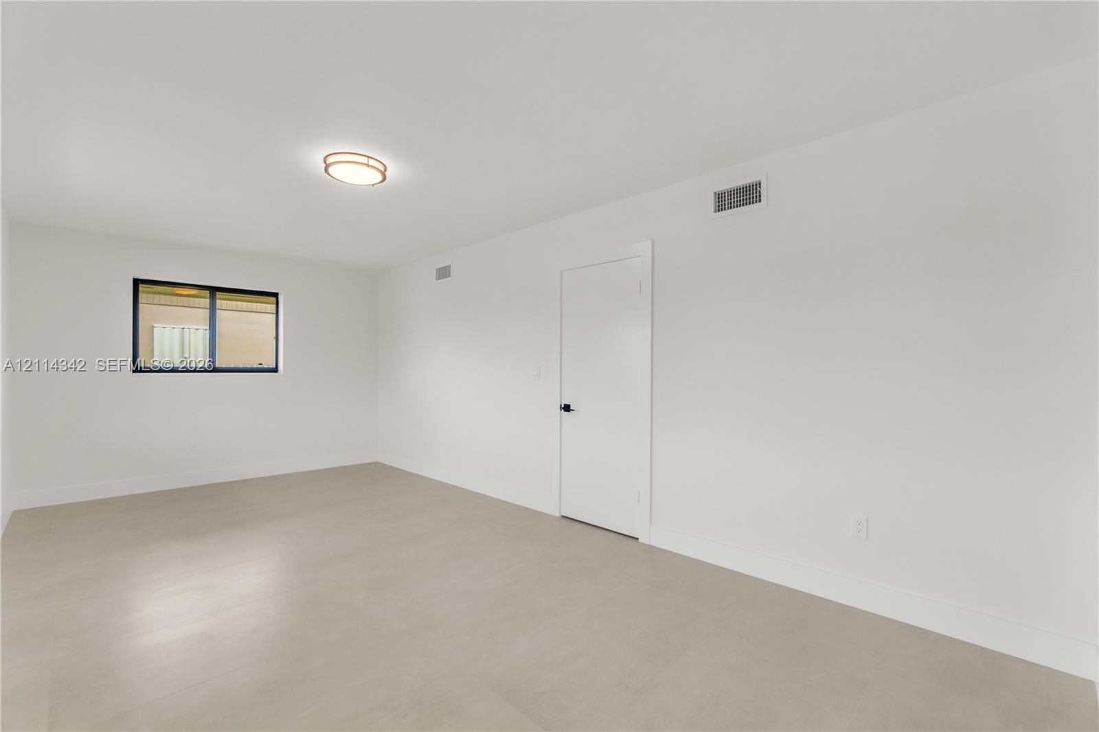 Neutral interior space featuring light-toned flooring, a single window with a black frame, and a circular flush-mount ceiling light fixture