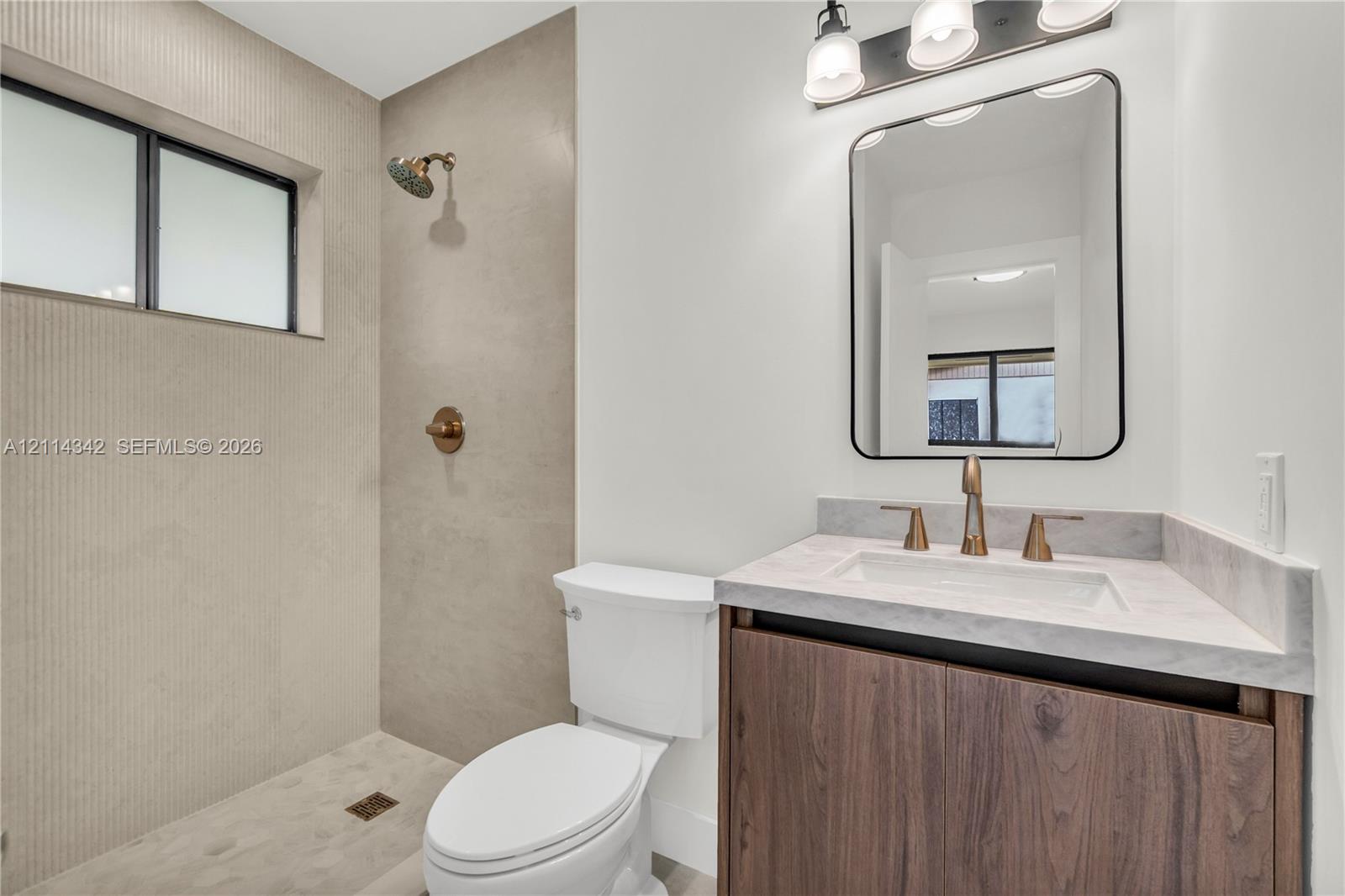 Bathroom featuring a wood-finish vanity with a stone countertop, wall-mounted rectangular mirror, three-light fixture, and a walk-in shower with large-format tile surround
