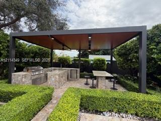 Outdoor kitchen featuring a stone-clad island, stainless steel grill, built-in refrigerator, and stone-topped bar under a covered pergola with integrated lighting