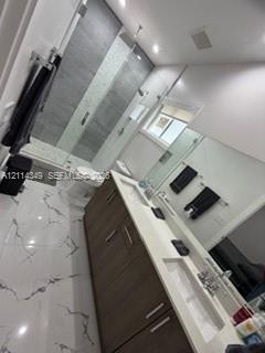 Dual-sink vanity with wood-finish cabinetry, white countertop, and integrated sinks, complemented by floor-to-ceiling tile in the walk-in glass-enclosed shower and large-format tile flooring