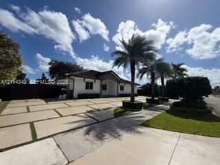 Single-story residence featuring a white stucco exterior, expansive paver driveway with turf accents, arched entryway, and mature palm trees Single-story residence featuring a white stucco exterior, expansive paver driveway with turf accents, arched entryway, and mature palm trees