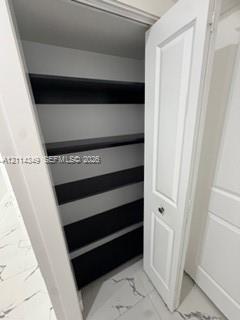 Storage closet featuring white paneled swing doors, multiple horizontal shelving tiers with dark-toned surfaces, and large-format tile flooring with veining detail