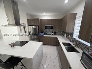 Kitchen featuring wood-finish cabinetry, quartz countertops, a central island with a built-in cooktop, stainless steel chimney-style range hood, and tile flooring Kitchen featuring wood-finish cabinetry, quartz countertops, a central island with a built-in cooktop, stainless steel chimney-style range hood, and tile flooring