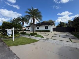 Single-story residence featuring a white exterior, dark shingle roof, expansive paver driveway with turf accents, and mature palm trees Single-story residence featuring a white exterior, dark shingle roof, expansive paver driveway with turf accents, and mature palm trees