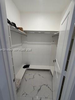 Walk-in storage space featuring white built-in shelving, integrated hanging rods, and large-format porcelain tile flooring with marble-finish veining