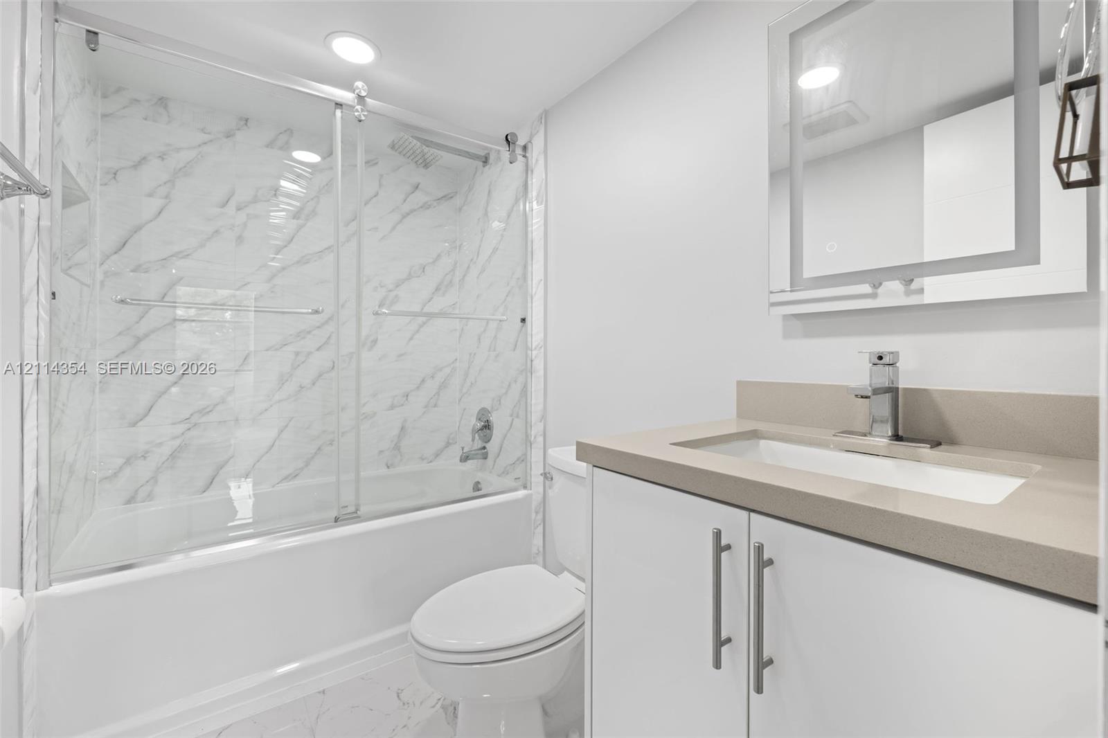 Bathroom featuring a tub-shower combination with floor-to-ceiling stone-pattern tile, a sliding glass door, a vanity with a solid-surface countertop and integrated sink, and a modern rectangular mirror with perimeter integrated lighting