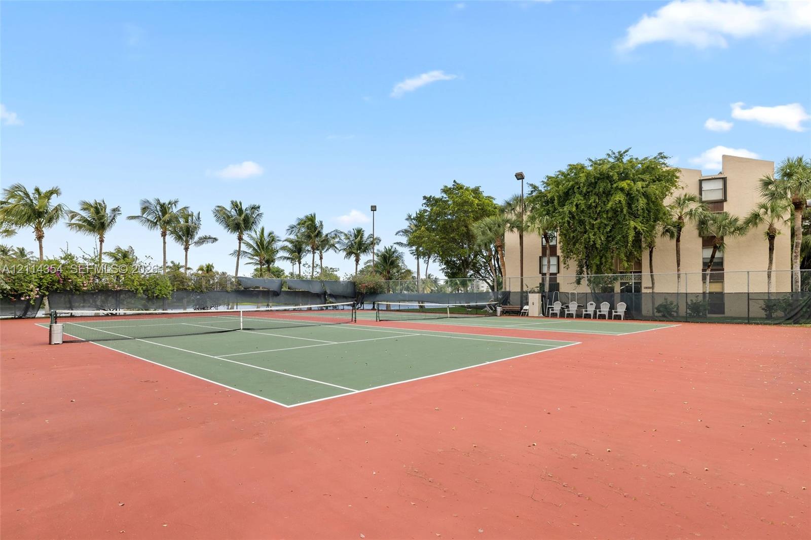 Hard-court tennis facility featuring perimeter chain-link fencing, dual courts, overhead lighting, and mature palm landscaping