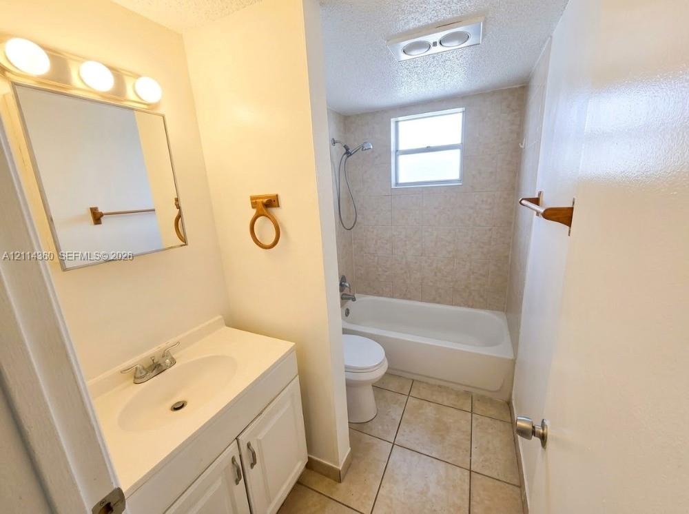 Bathroom featuring a white vanity with integrated sink, three-light wall-mounted fixture, tiled flooring, and a shower-tub combination with wall tiling