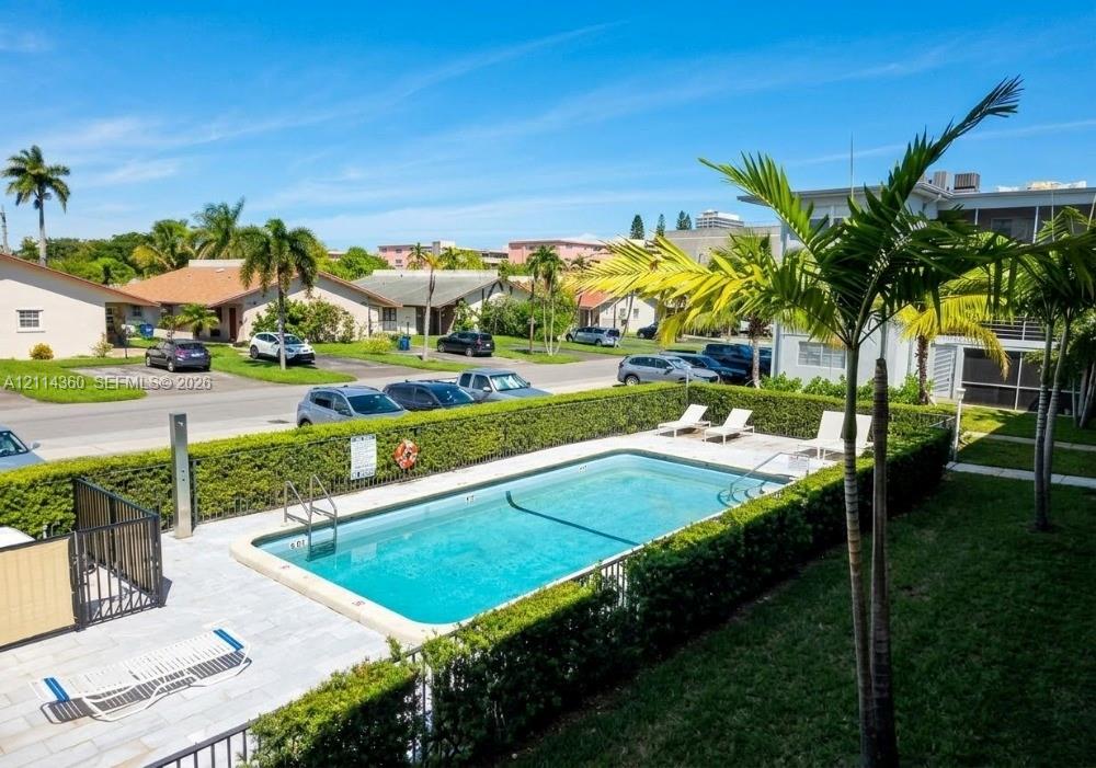 Swimming pool with stone-tiled decking, enclosed by a manicured hedge, palm trees, and metal fencing