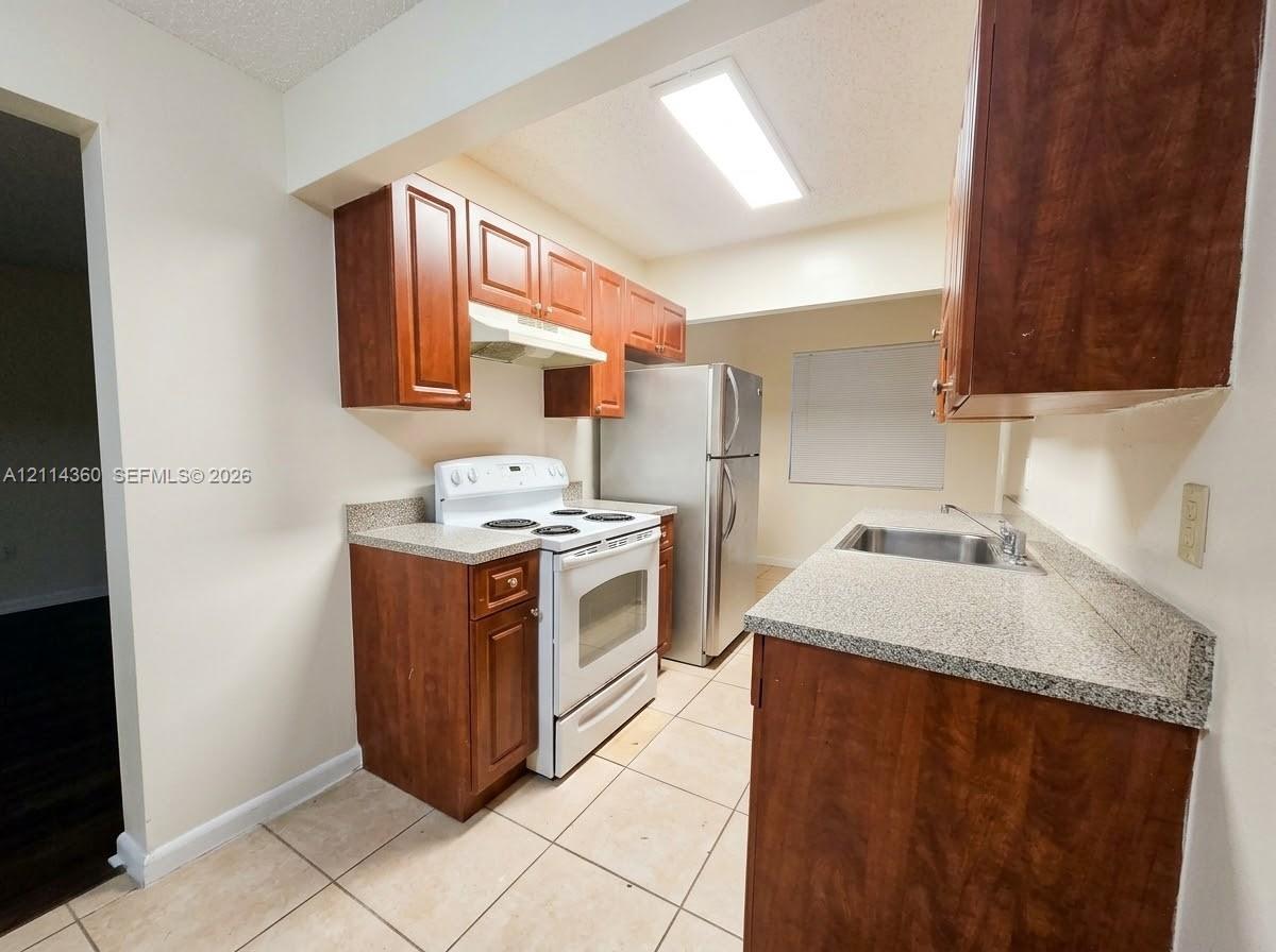 Kitchen featuring wood cabinetry, stone-composite countertops, stainless steel refrigerator, white freestanding range, and tile flooring Kitchen featuring wood cabinetry, stone-composite countertops, stainless steel refrigerator, white freestanding range, and tile flooring