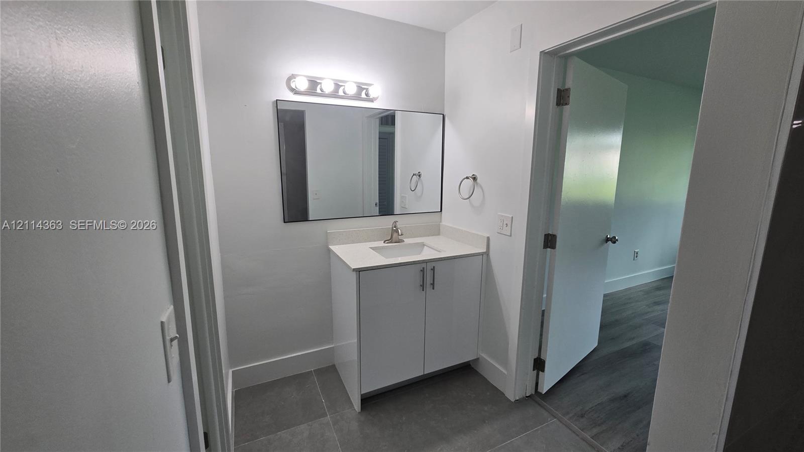 White vanity with stone-composite countertop, undermount sink, rectangular wall mirror, four-bulb vanity light fixture, and large-format tile flooring