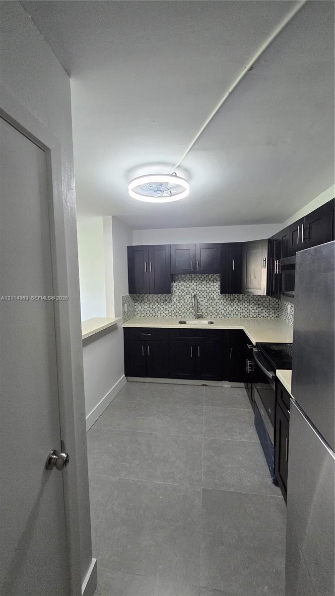 Kitchen featuring dark cabinetry, solid-surface countertops, mosaic tile backsplash, stainless steel appliances, and large-format floor tile