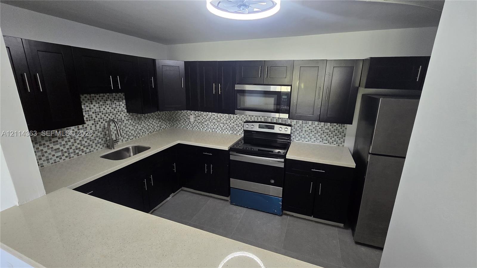 Kitchen featuring dark wood-finish cabinetry, light-toned stone countertops, mosaic tile backsplash, stainless steel appliances, and a pass-through breakfast bar