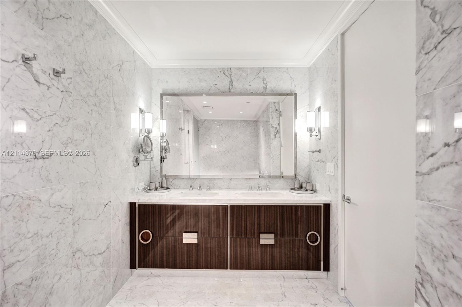 Double vanity with a stone countertop, dark wood-finish cabinetry, a large rectangular mirror, and wall-mounted metal light fixtures, all encased in full-height stone wall cladding