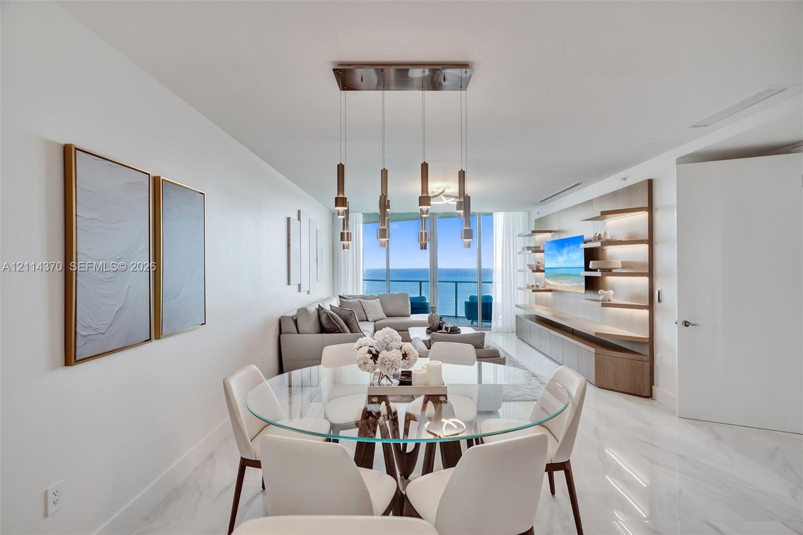 Large-format polished stone flooring, custom integrated cabinetry with illuminated shelving, multi-pendant rectangular lighting fixture, floor-to-ceiling glass paneling, and ocean scenery