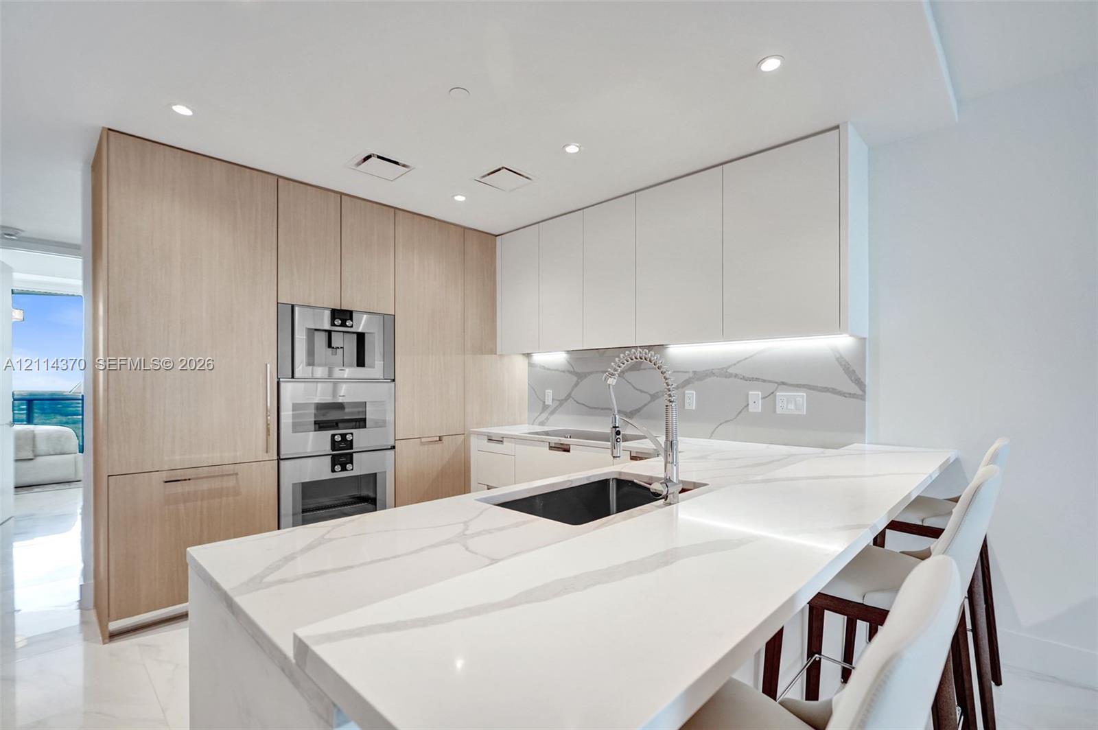 Kitchen featuring light-tone flat-panel cabinetry, white stone-finish waterfall island, integrated stainless steel double oven, and matching stone-finish backsplash with under-cabinet lighting