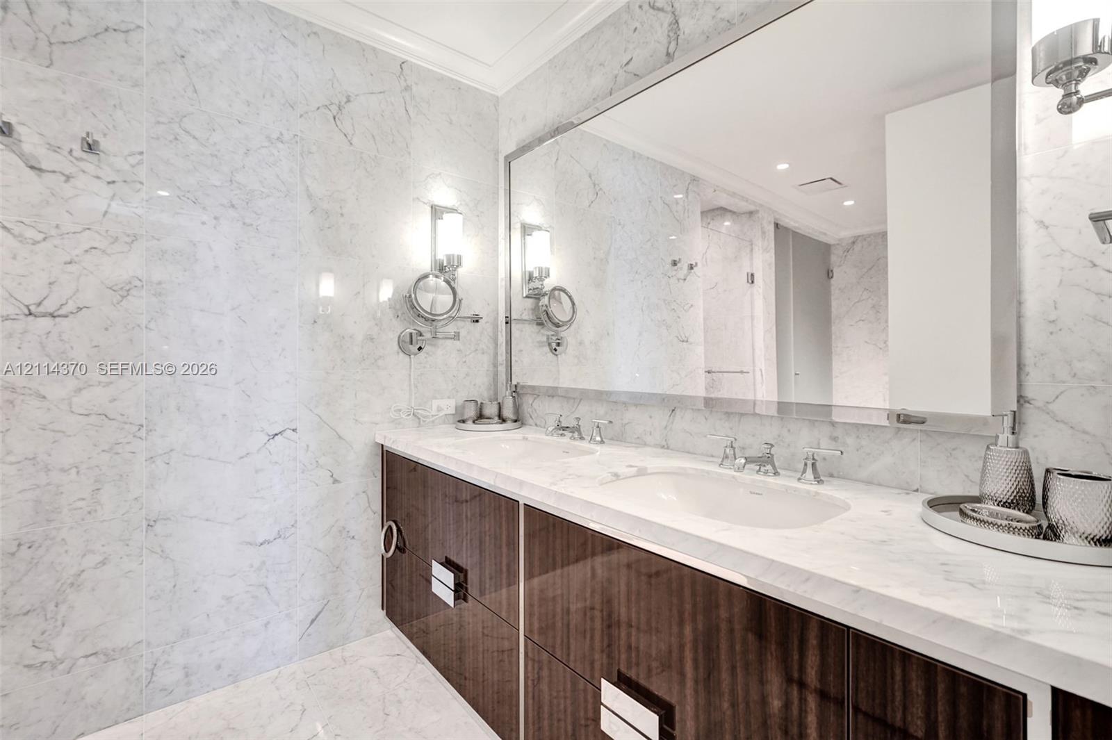 Dual-sink vanity with high-gloss cabinetry, stone-surfaced countertops, full-width wall mirror, wall-mounted lighting fixtures, and floor-to-ceiling stone wall tiling