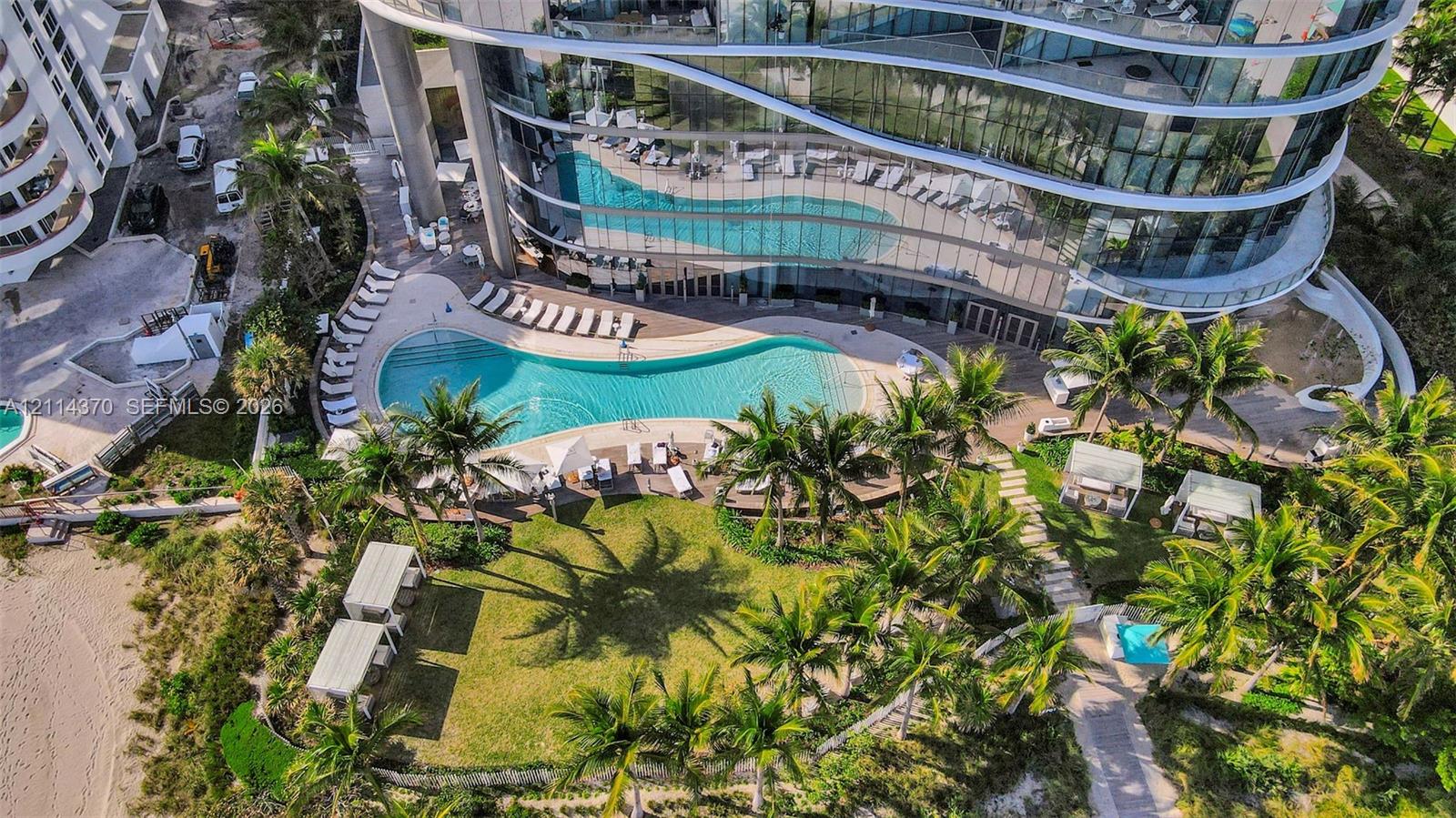 Curved high-rise building with floor-to-ceiling glass windows, expansive curved swimming pool, manicured lawn, palm-lined landscaping, and paved walkways