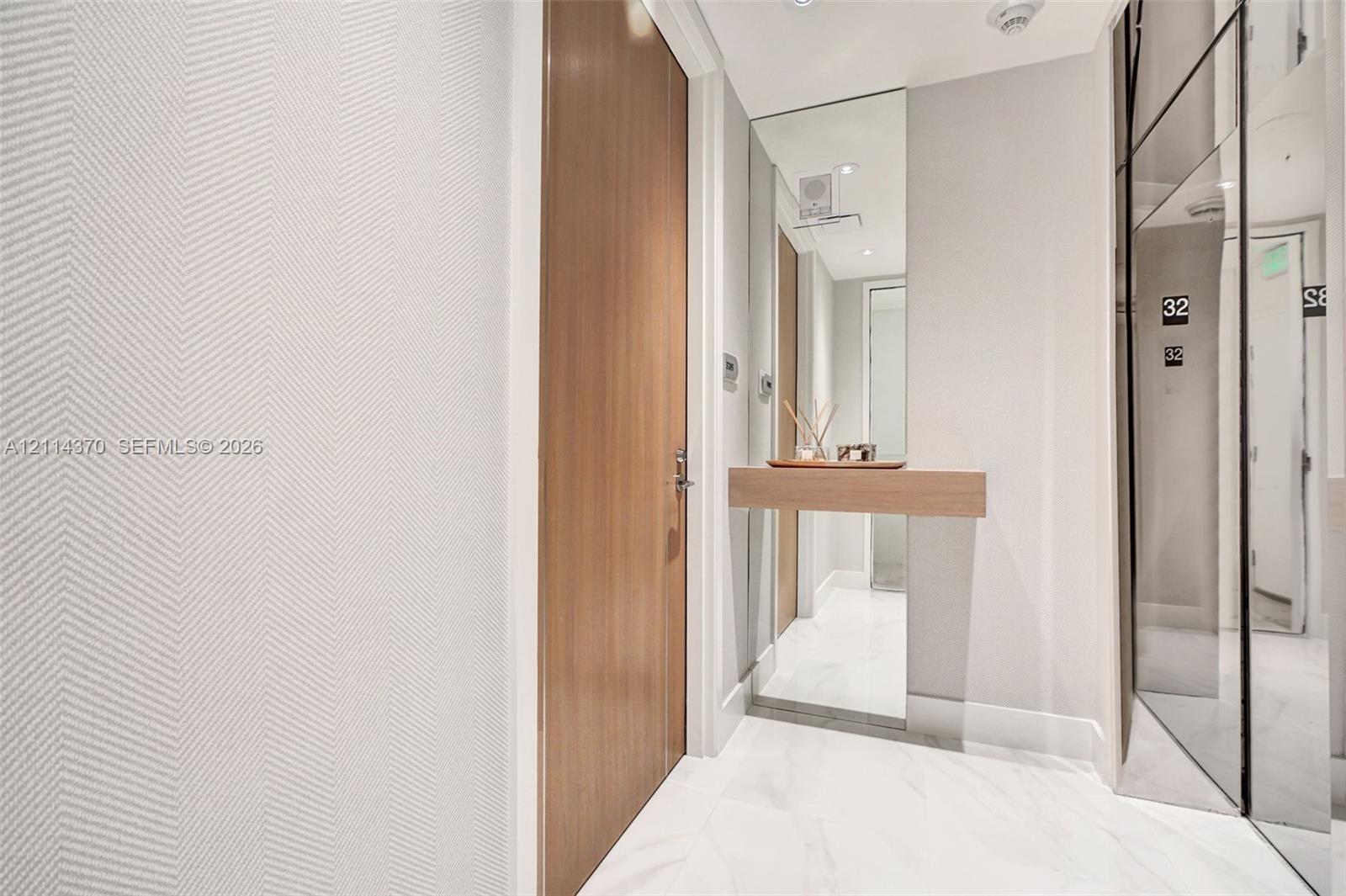Textured chevron-patterned wall covering, full-height mirrored wall panel, stone-finish flooring, wood-veneer door, and floating wood-finish console shelf