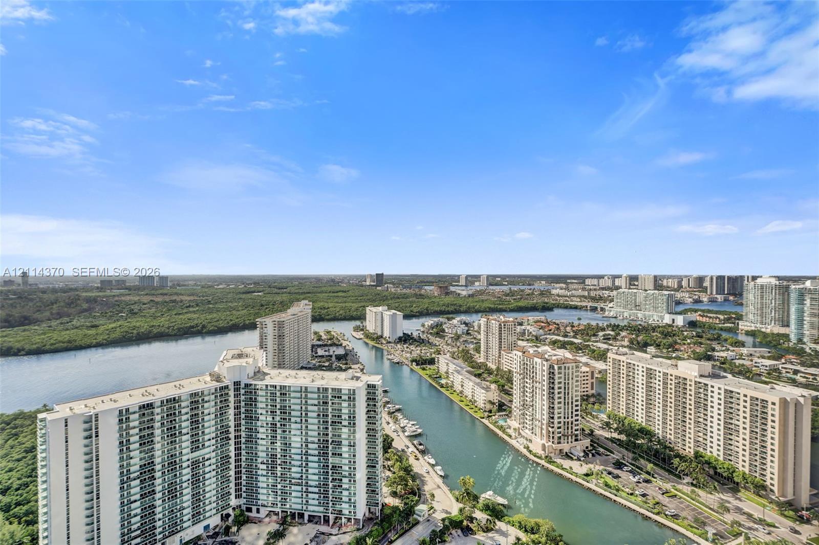 Panoramic coastal landscape featuring a waterway, high-rise residential buildings, and dense natural foliage under a clear sky