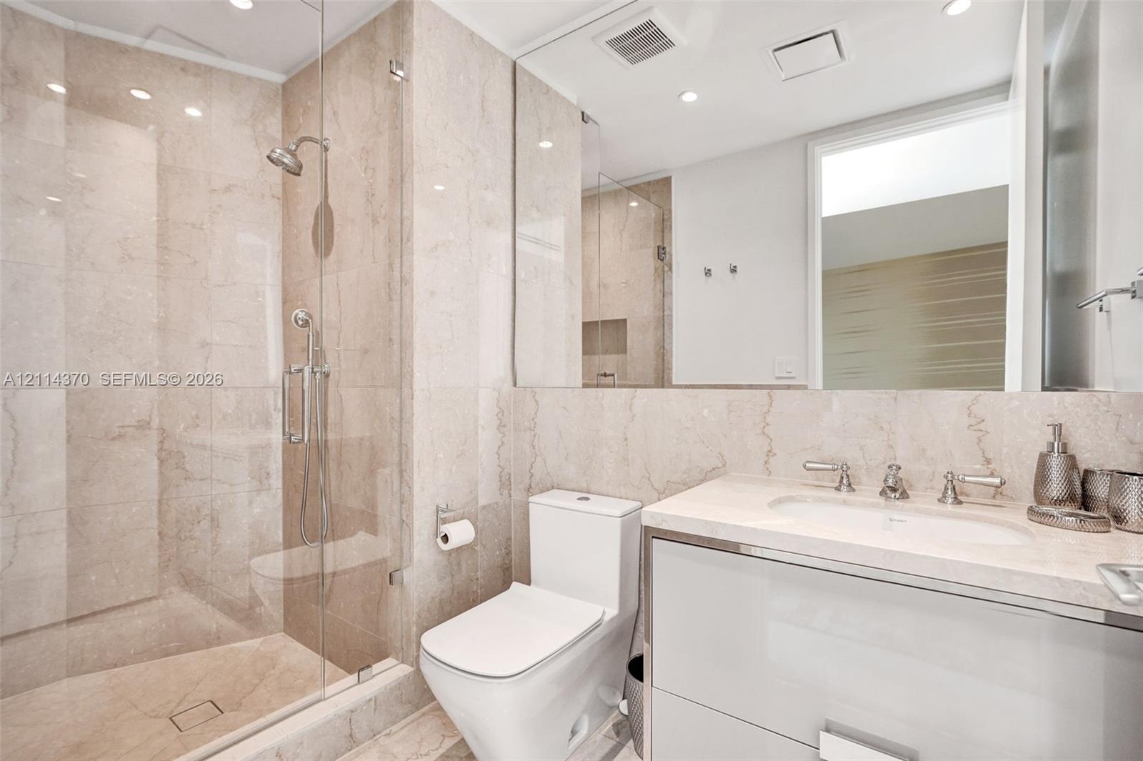 Full-height stone wall tiling, glass-enclosed shower, stone vanity countertop with integrated sink, and wall-to-wall mirror