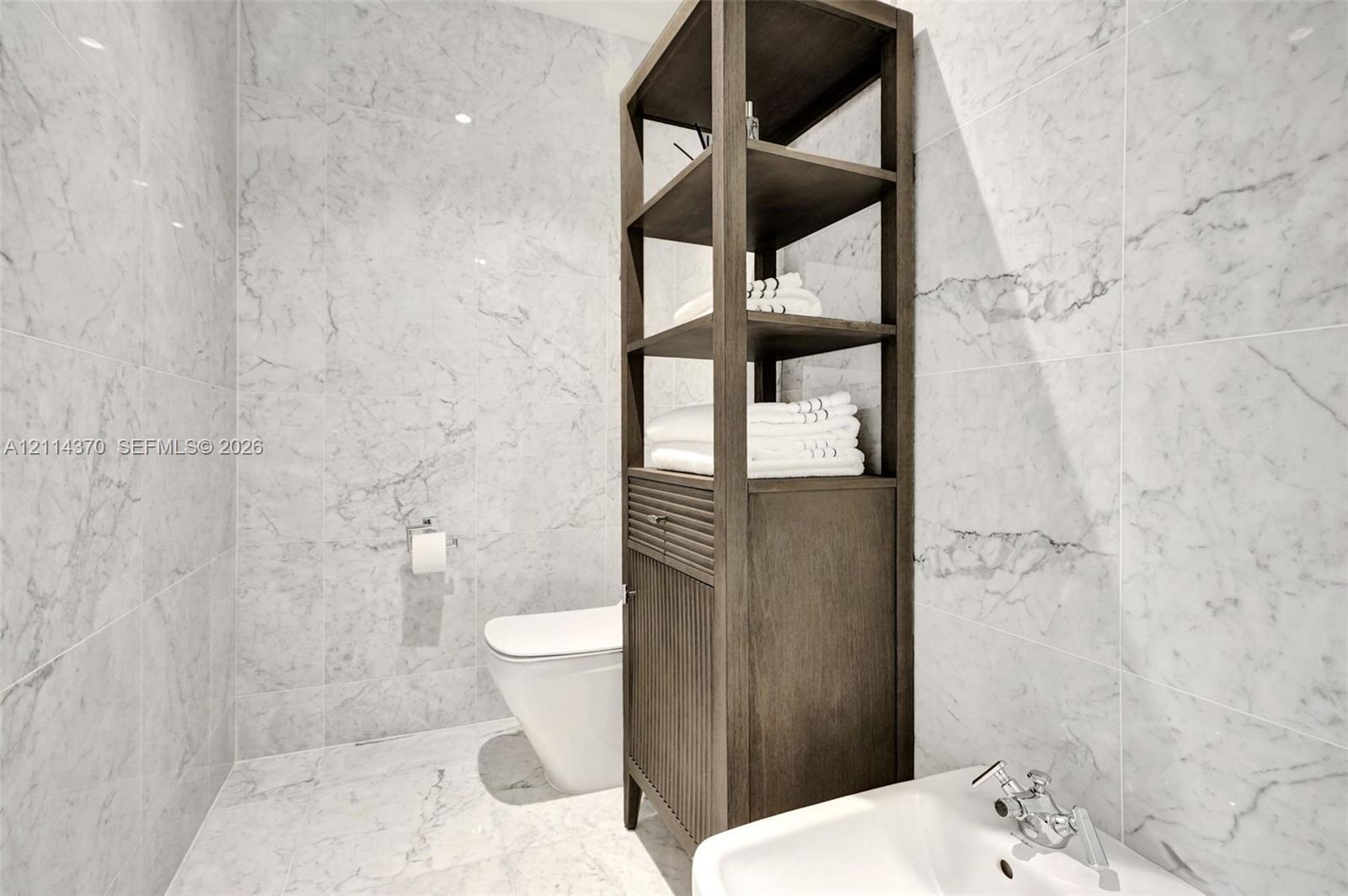 Full-height stone wall tiling, white wall-mounted toilet, custom dark wood built-in shelving unit with lower cabinetry, and wall-mounted basin