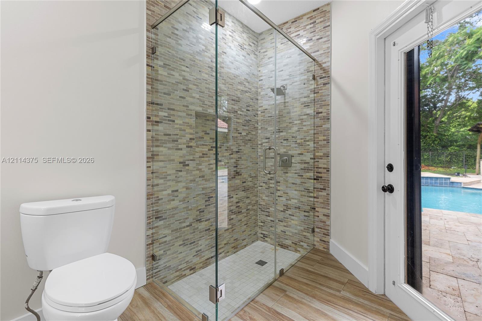 Walk-in shower featuring a frameless glass enclosure, recessed wall niche, and textured multi-tonal tile surround, complemented by wood-finish flooring and direct external access