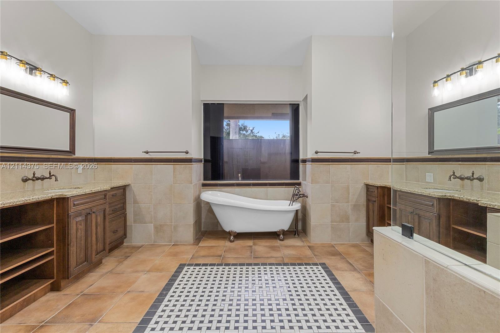 Freestanding clawfoot tub, dual wooden vanity cabinetry with stone countertops, decorative tile floor inlay, and wall-mounted vanity lighting