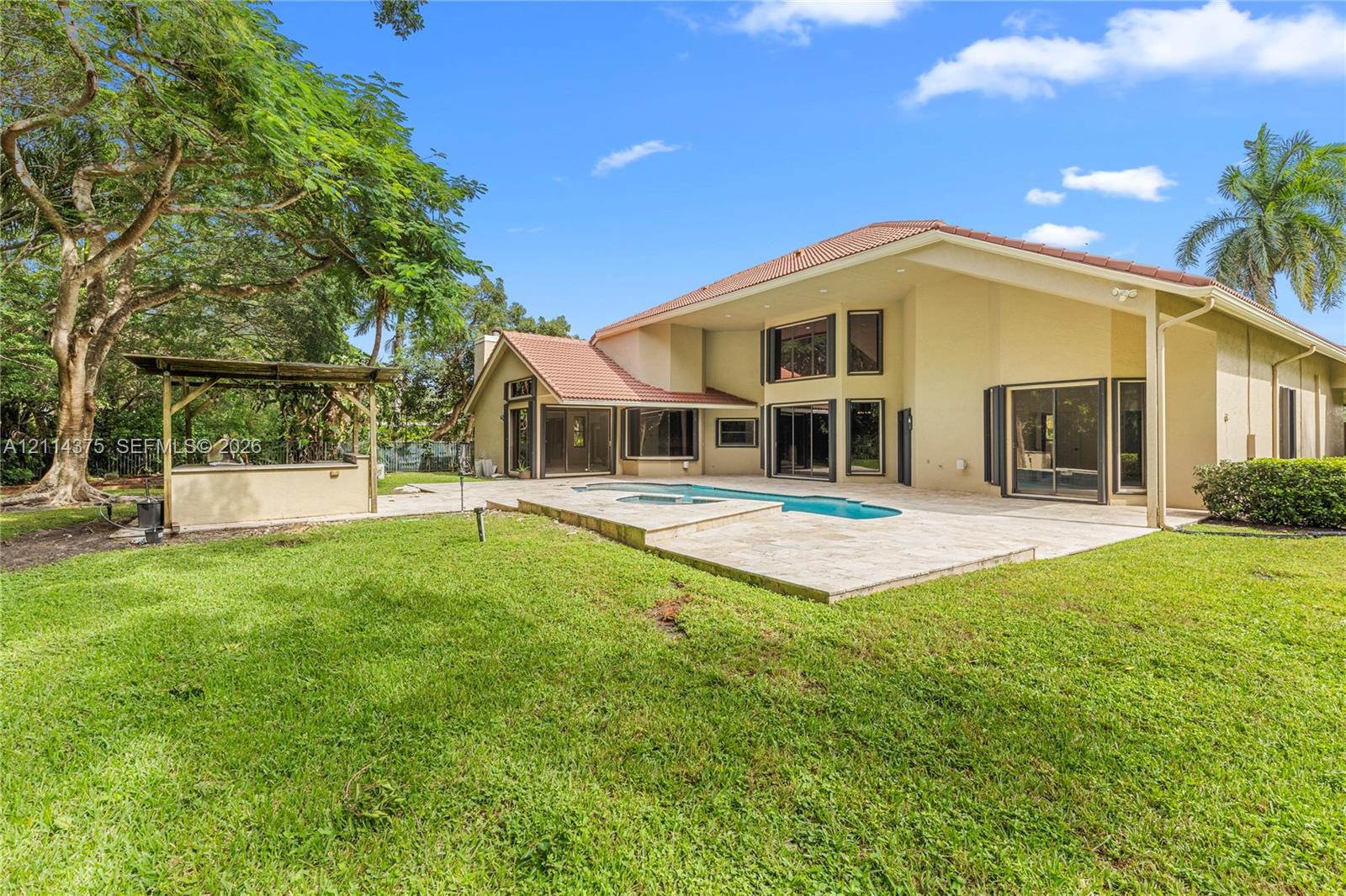 Rear exterior featuring a swimming pool, stone-paver patio, built-in outdoor bar, and manicured lawn with mature trees