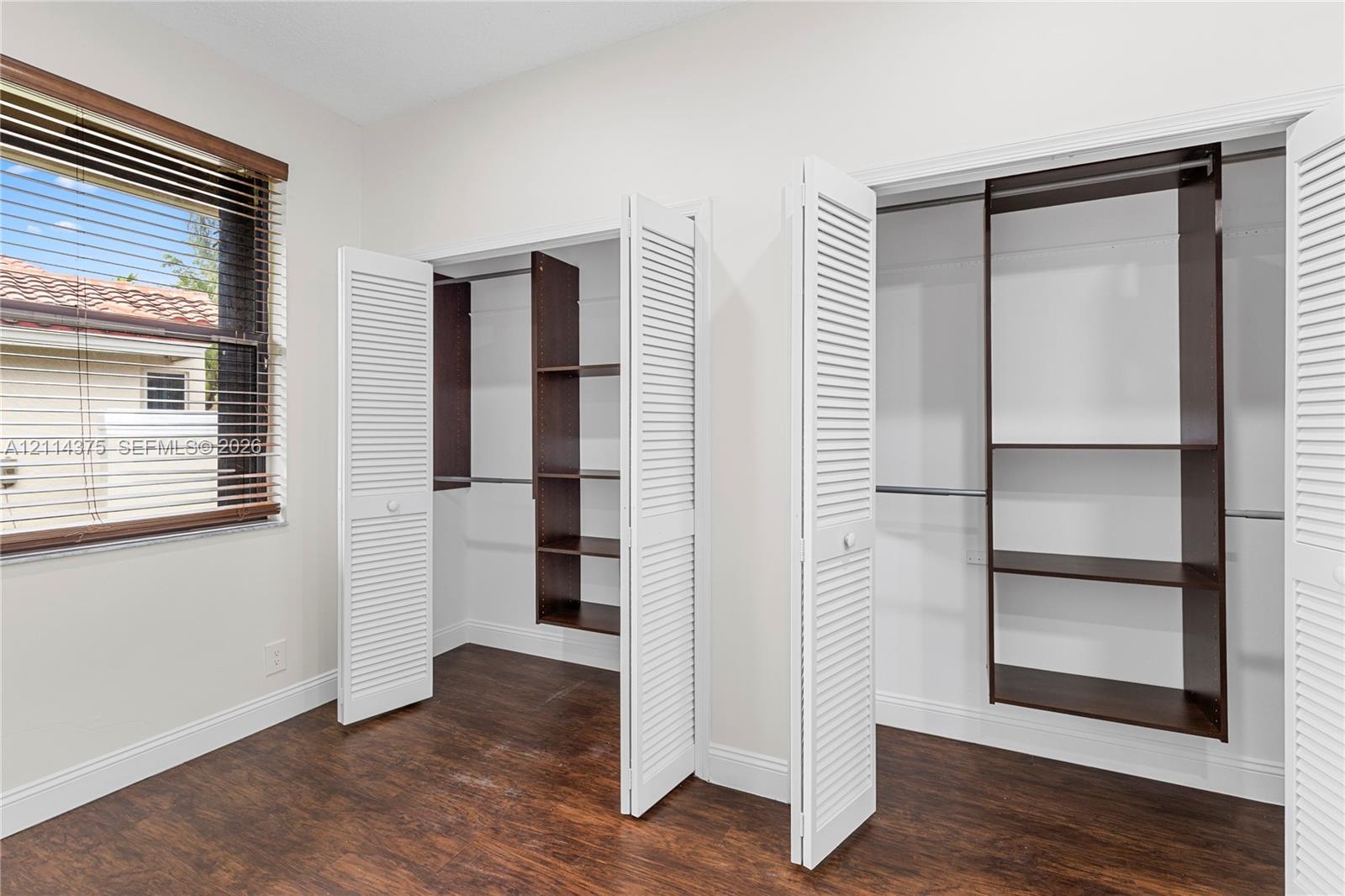 Wood-finish flooring, dual closets with white louvered doors, and built-in dark wood shelving units with integrated hanging rods