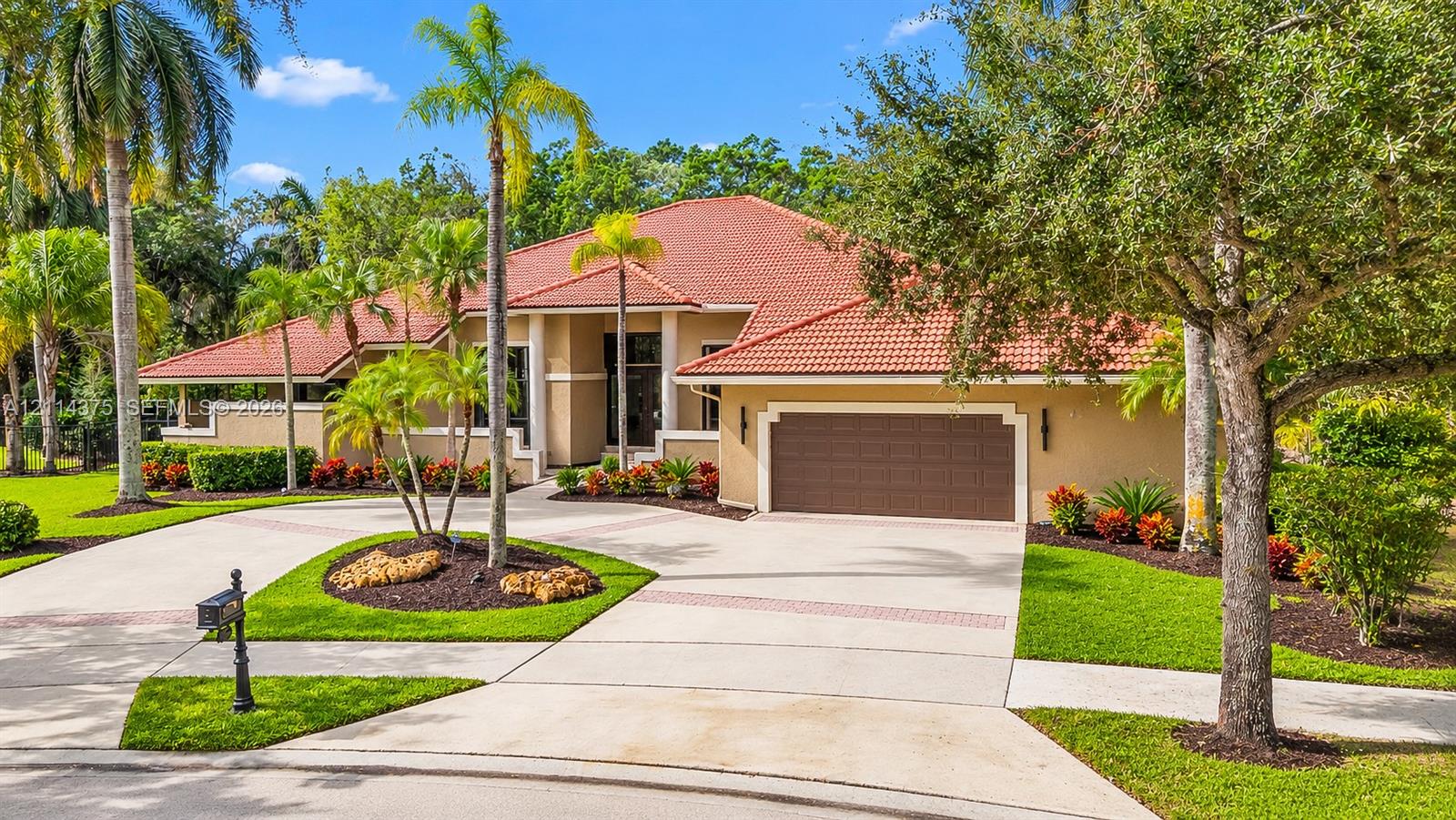 Stucco exterior featuring a terracotta tile roof, an oversized multi-car garage, and a paver-accented concrete driveway situated within a landscaped lot with mature palm trees