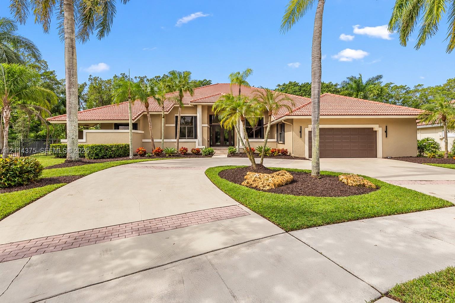 Stucco exterior featuring a terracotta-tile roof, expansive concrete driveway with brick inlay, and mature palm landscaping