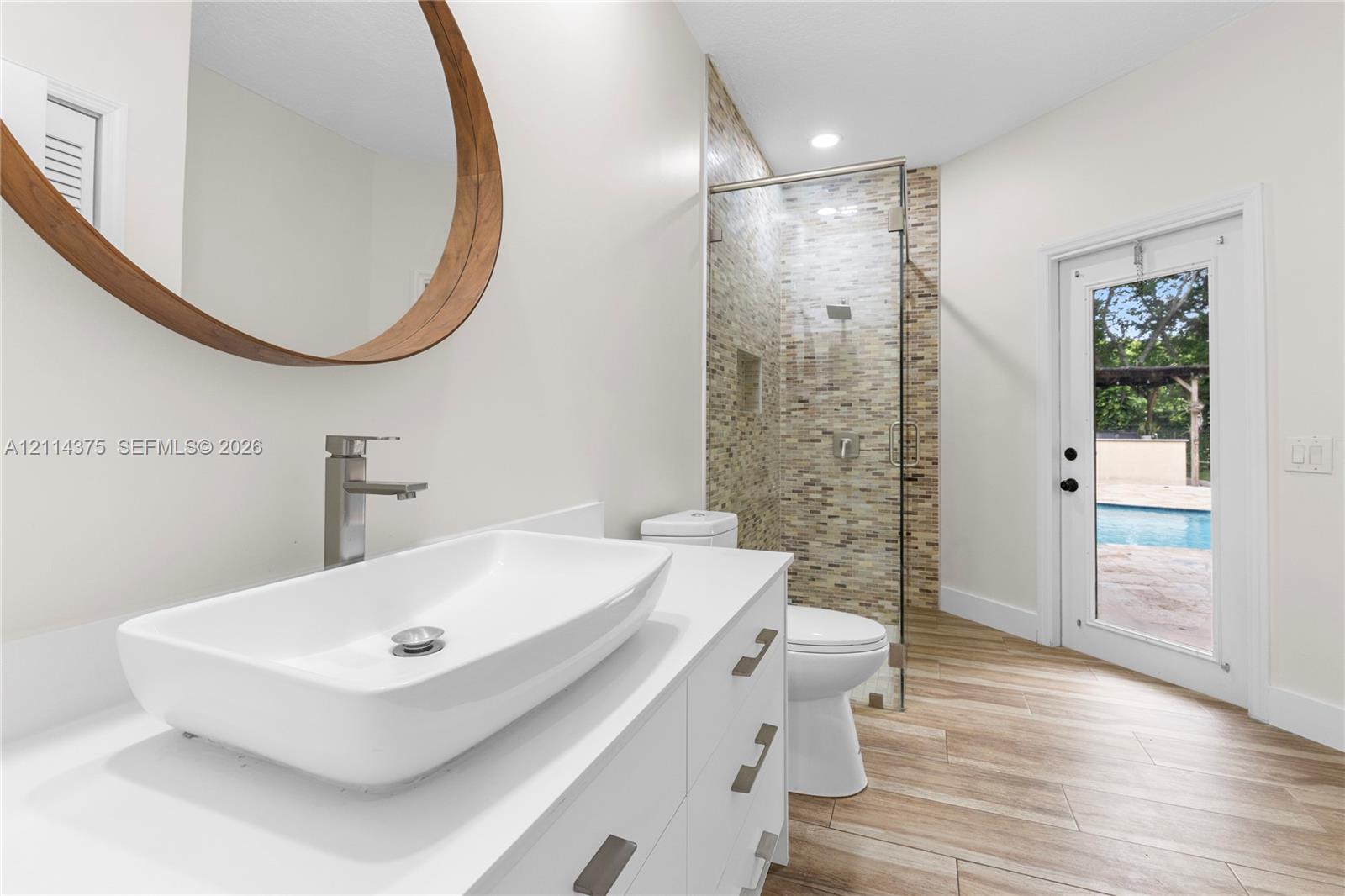 Bathroom featuring a rectangular vessel sink with a chrome faucet, wood-framed circular mirror, walk-in shower with stone-tile surround, wood-finish flooring, and a glass-paneled exterior door