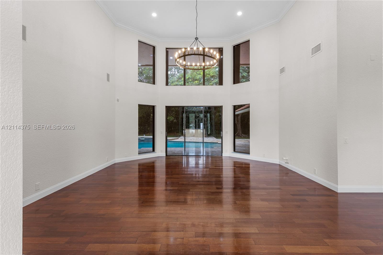Expansive room featuring high ceilings, wood-finish flooring, floor-to-ceiling windows, a multi-tier circular chandelier, and sliding glass doors