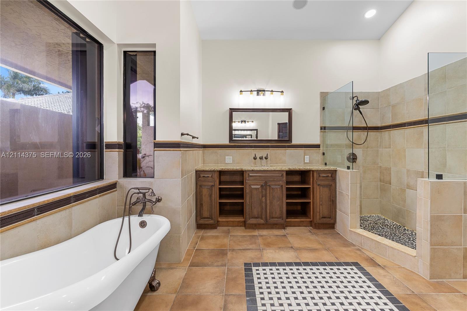 Freestanding soaking tub with dark metal faucet, dual-sink wooden vanity with stone countertop, tiled walk-in shower with glass enclosure, and patterned tile floor inlay