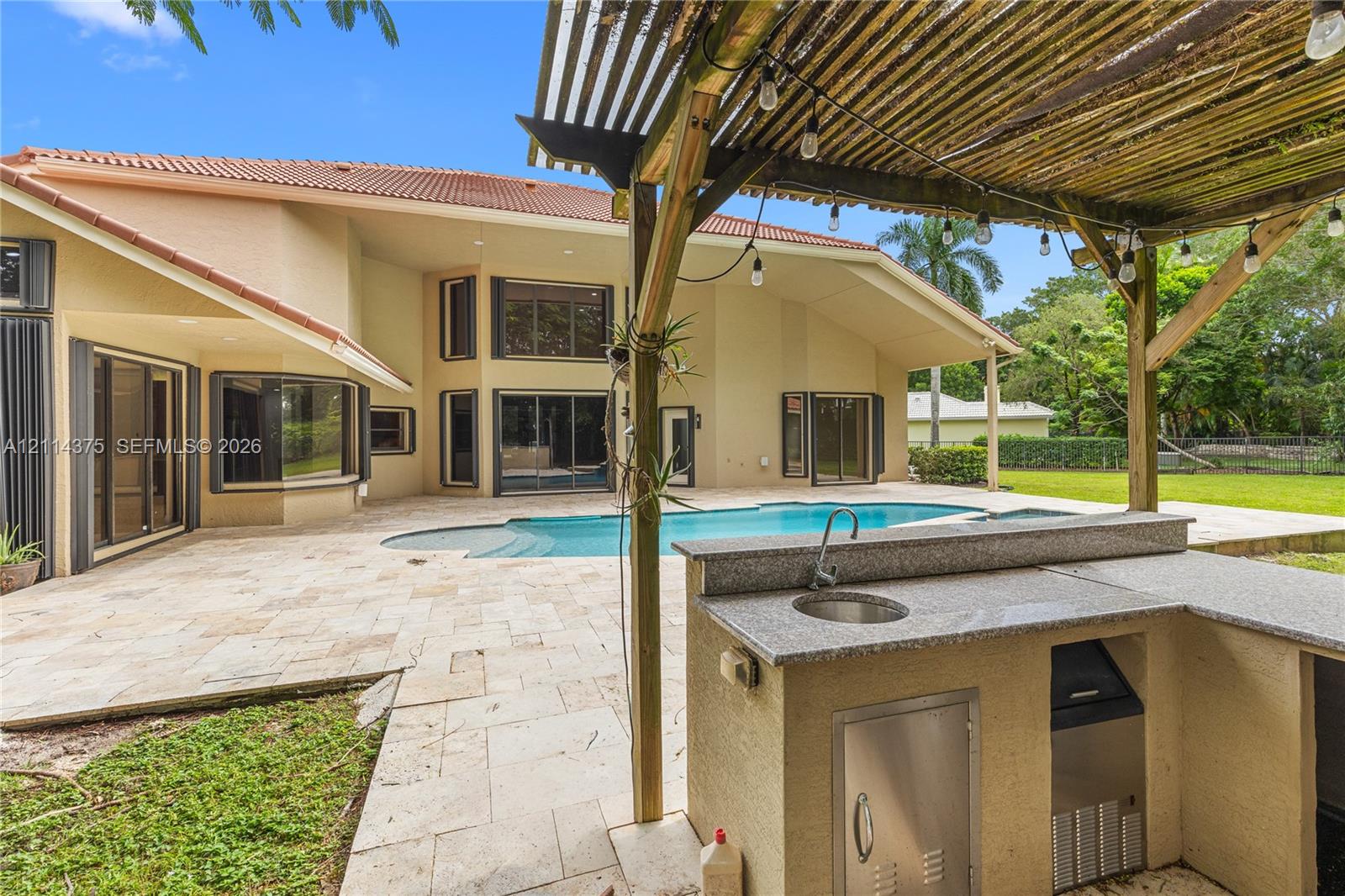 Outdoor kitchen featuring stone-finish counters, integrated sink, and stainless-steel cabinetry under a wooden pergola, situated on a stone-paver patio with a custom pool