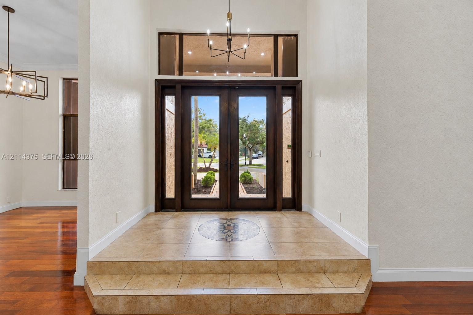Entryway featuring double glass-paneled doors, decorative tile inlay, stepped tile flooring, transom window, and multi-arm chandelier