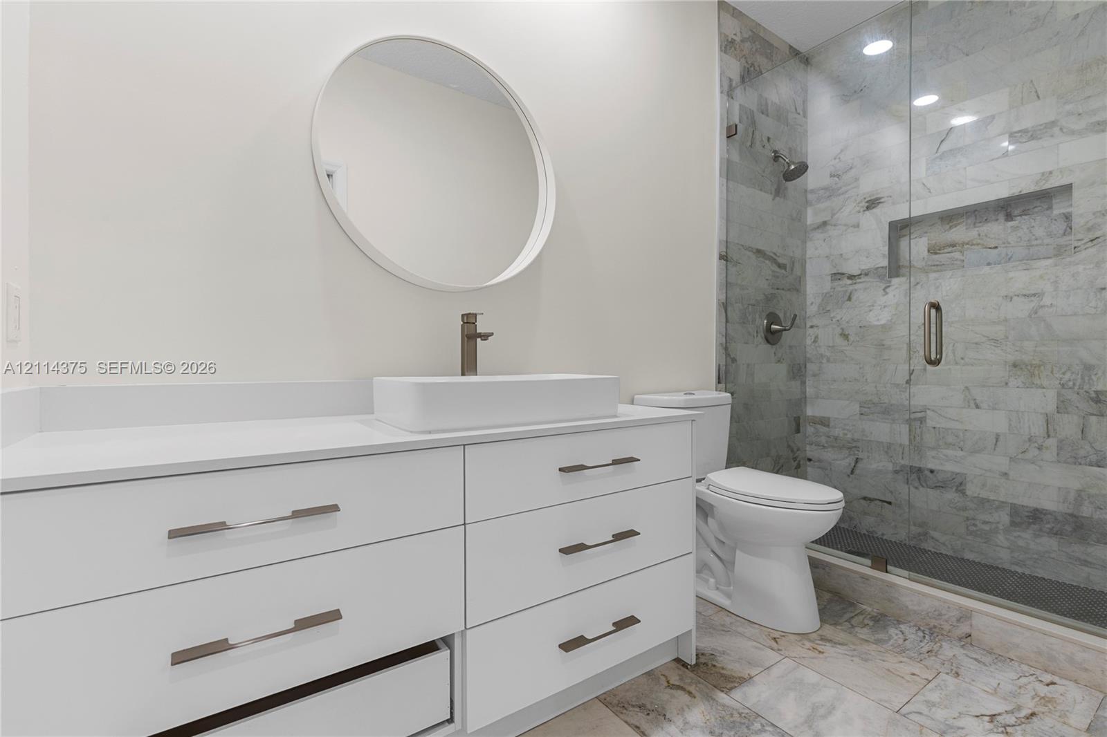 White cabinetry vanity with rectangular vessel sink, frameless glass shower enclosure with stone-tile surround, built-in shower niche, and round wall-mounted mirror