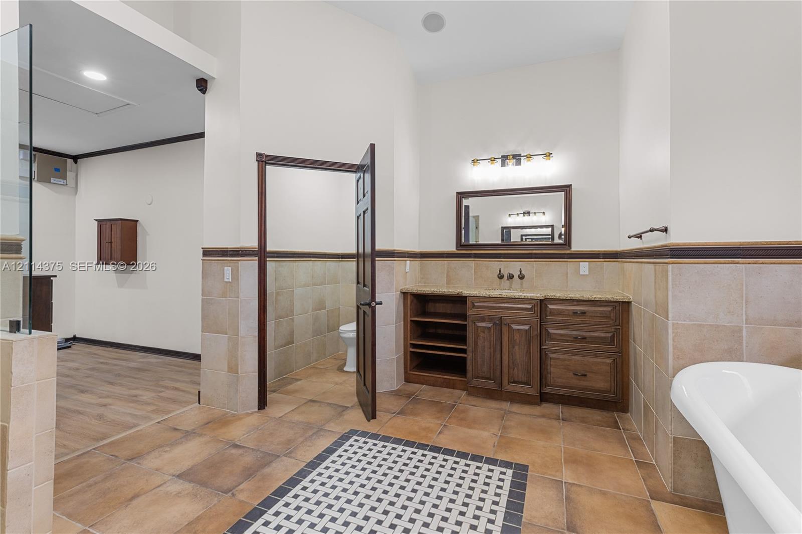 Bathroom featuring a wood-tone vanity with stone countertop, tile wainscoting, a wall-mounted framed mirror, a soaking tub, and a decorative tile inlay floor
