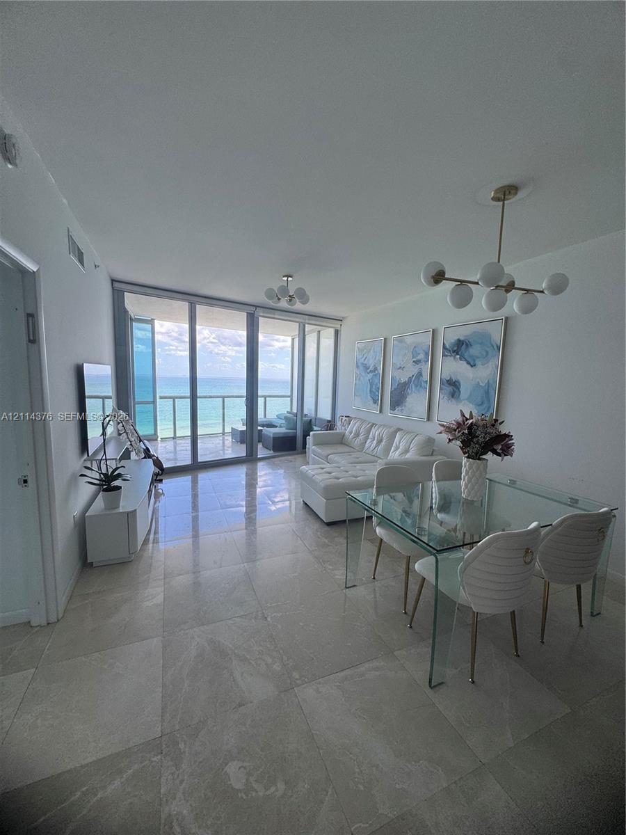 Open-concept living space featuring floor-to-ceiling glass panels, large-format tile flooring, multi-globe brass-finish chandeliers, and a private balcony with ocean frontage