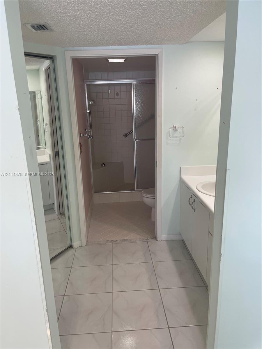 Bathroom featuring large-format ceramic tile flooring, white vanity cabinet with a solid surface countertop, integrated sink basin, and a walk-in shower with glass sliding doors