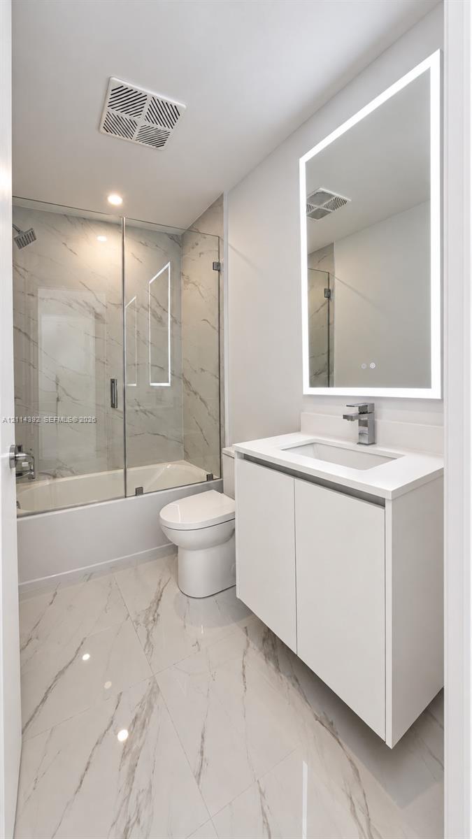 Bathroom featuring a floating vanity with a white countertop, wall-mounted illuminated mirror, porcelain tile flooring, and a glass-enclosed shower with floor-to-ceiling tile surround
