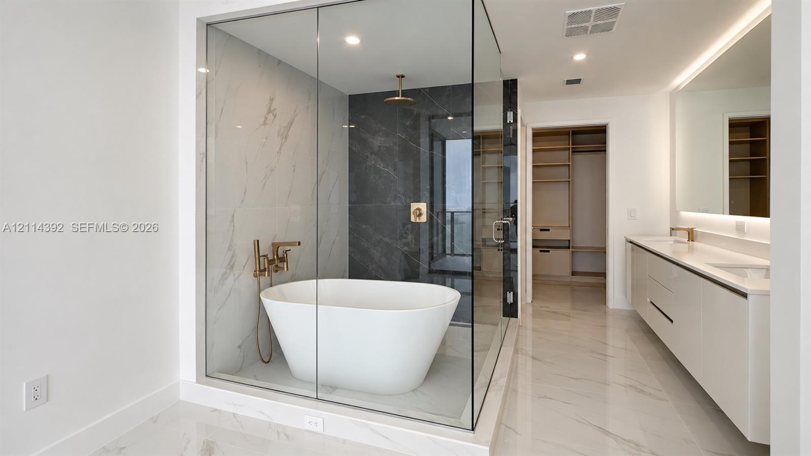 Glass-enclosed wet room featuring a freestanding soaking tub, dual-tone tile surround, and brushed gold fixtures, complemented by a dual-sink vanity with under-cabinet lighting and a walk-in dressing area with built-in shelving Glass-enclosed wet room featuring a freestanding soaking tub, dual-tone tile surround, and brushed gold fixtures, complemented by a dual-sink vanity with under-cabinet lighting and a walk-in dressing area with built-in shelving