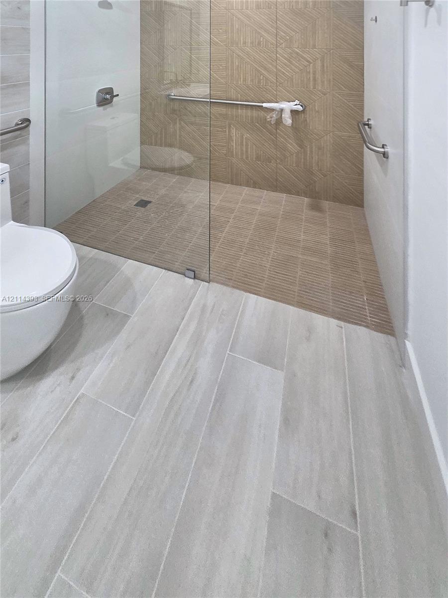 Walk-in shower featuring a frameless glass enclosure, geometric-patterned tile wall accents, wood-finish floor tiling, and wall-mounted metal support bars