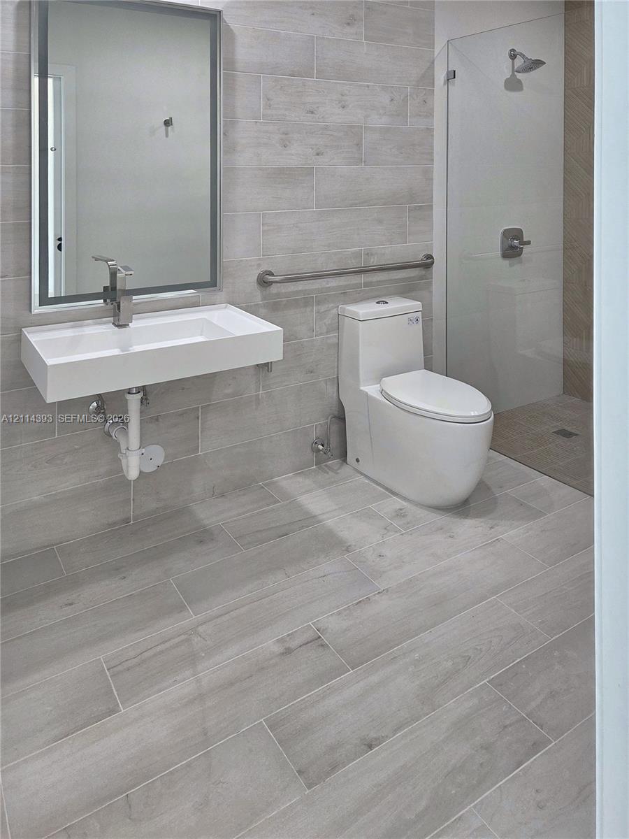 Wall-mounted rectangular basin with a single-handle faucet, full-height tile wall surfacing, walk-in glass shower enclosure, elongated one-piece toilet, and wood-finish tile flooring